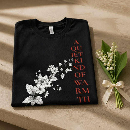Eternal Light black unisex t-shirt neatly folded on a textured stone table. The print showcases a white botanical floral sketch and vertical crimson red typography reading A Quiet Kind of Warmth, beautifully styled next to a delicate bouquet of white Lily of the Valley flowers tied with a cream silk ribbon.