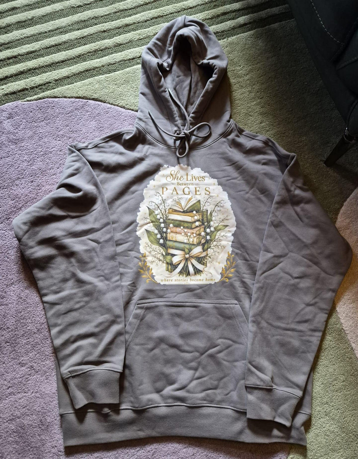 An extra-heavyweight unisex hoodie in a rich olive green colour, shown lying flat on a geometric rug. The chest features an elegant, hand-drawn dark academia graphic showcasing a stack of vintage books intertwined with delicate white Lily of the Valley blossoms. Ornate script reads, "She Lives Between Pages Where Stories Become Home."