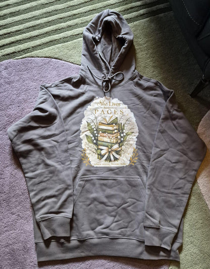 An extra-heavyweight unisex hoodie in a rich olive green colour, shown lying flat on a geometric rug. The chest features an elegant, hand-drawn dark academia graphic showcasing a stack of vintage books intertwined with delicate white Lily of the Valley blossoms. Ornate script reads, "She Lives Between Pages Where Stories Become Home."