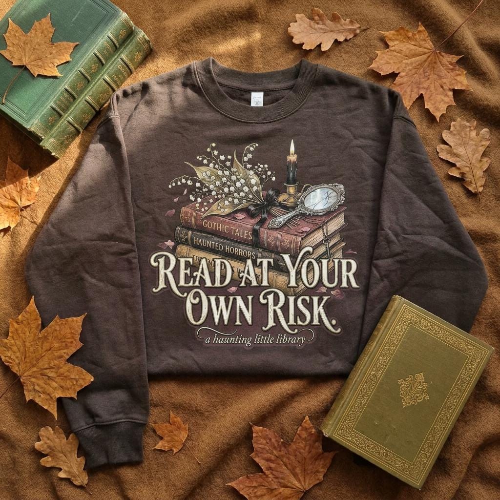 A premium 370GSM heavyweight unisex sweatshirt in a unique charcoal espresso brown, folded on a blanket. The front displays a detailed dark academia design featuring a stack of vintage books titled "Gothic Tales." A burning black candle, delicate lily of the valley blossoms, and a vintage cracked mirror rest atop the books. Cream text boldly reads "READ AT YOUR OWN RISK." The cozy arrangement is framed by rustic, dried autumn oak leaves and antique library volumes.