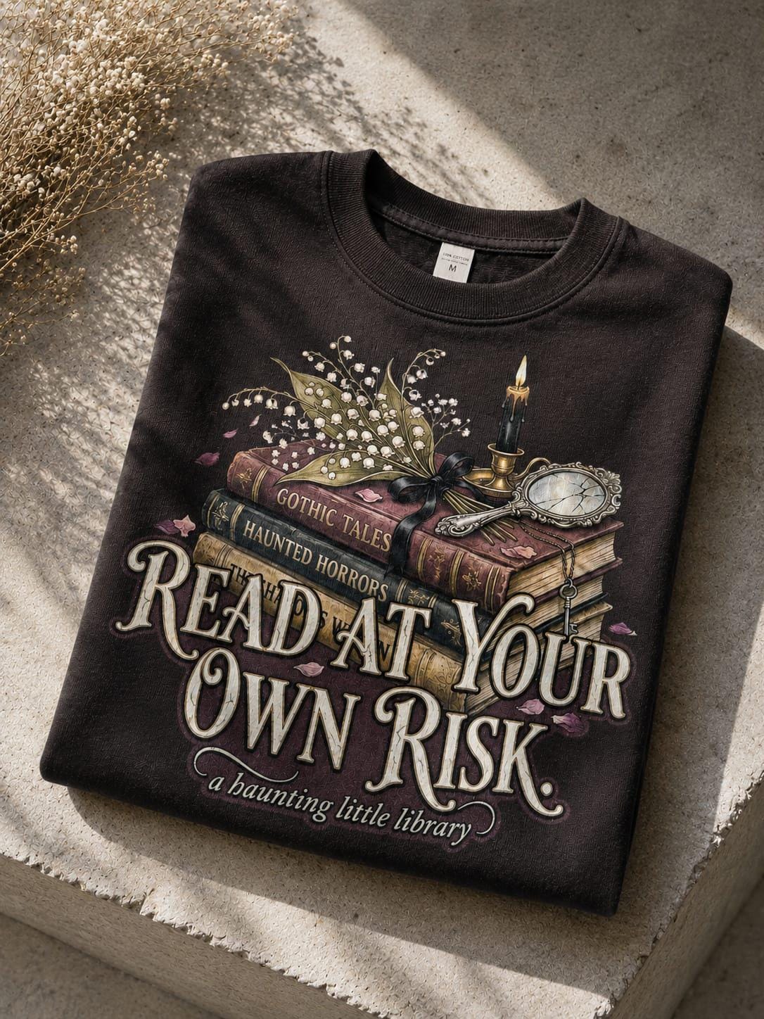 A premium 370GSM heavyweight unisex sweatshirt in a unique charcoal espresso brown, neatly folded on a beige stone shelf. The front displays a detailed dark academia design featuring a stack of vintage books titled "Gothic Tales." A burning black candle, delicate lily of the valley blossoms, and a vintage cracked mirror rest atop the books. Cream text boldly reads "READ AT YOUR OWN RISK." The cozy arrangement is framed by rustic, dried florals.