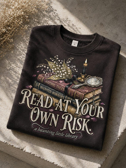 A premium 370GSM heavyweight unisex sweatshirt in a unique charcoal espresso brown, neatly folded on a beige stone shelf. The front displays a detailed dark academia design featuring a stack of vintage books titled "Gothic Tales." A burning black candle, delicate lily of the valley blossoms, and a vintage cracked mirror rest atop the books. Cream text boldly reads "READ AT YOUR OWN RISK." The cozy arrangement is framed by rustic, dried florals.