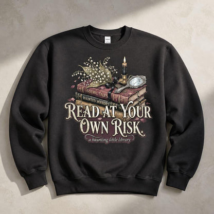 A premium 370GSM heavyweight sweatshirt in an earthy charcoal espresso tone, laid completely flat to showcase its relaxed, oversized unisex silhouette. The center chest displays an intricate cream graphic of an antique book stack intertwined with delicate white lily of the valley petals. Large, stylized text reads "READ AT YOUR OWN RISK" above a script banner that states "a haunting little library." The fabric exhibits a thick, structured drape perfect for cozy autumn styling.