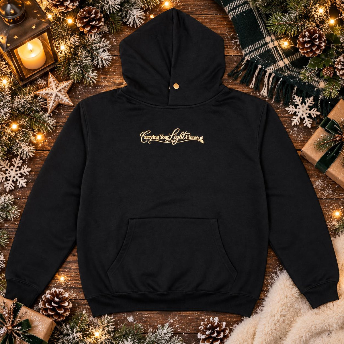 Black embroidered hoodie with gold Carrying Your Light Home design styled in a cosy Christmas setting.