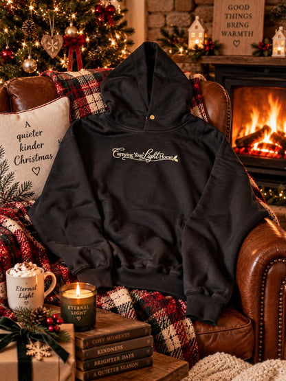 Black embroidered hoodie with gold Carrying Your Light Home design styled beside a warm fireplace.