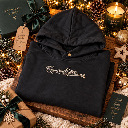 Black Carrying Your Light Home embroidered hoodie displayed in a cosy winter setting with festive greenery.