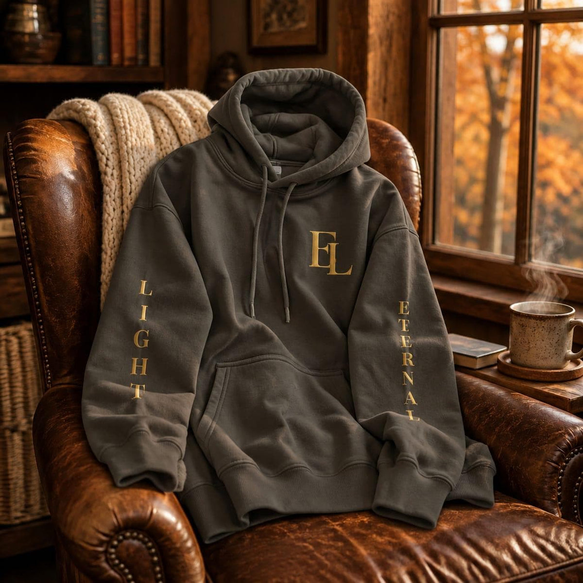 Charcoal green Eternal Light hoodie resting on a brown leather armchair beside an autumn window and a steaming mug. Gold lettering is visible on the chest and sleeves.
