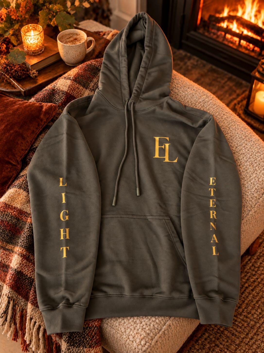 Charcoal green hoodie laid flat on a knitted throw by the fire, with a gold EL monogram on the chest and LIGHT and ETERNAL down the sleeves.