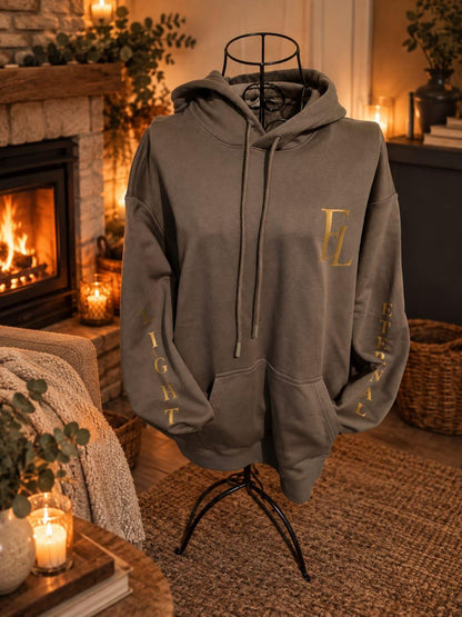 Charcoal green Eternal Light hoodie displayed on a dress form beside a lit fireplace, showing its gold chest monogram and sleeve lettering.