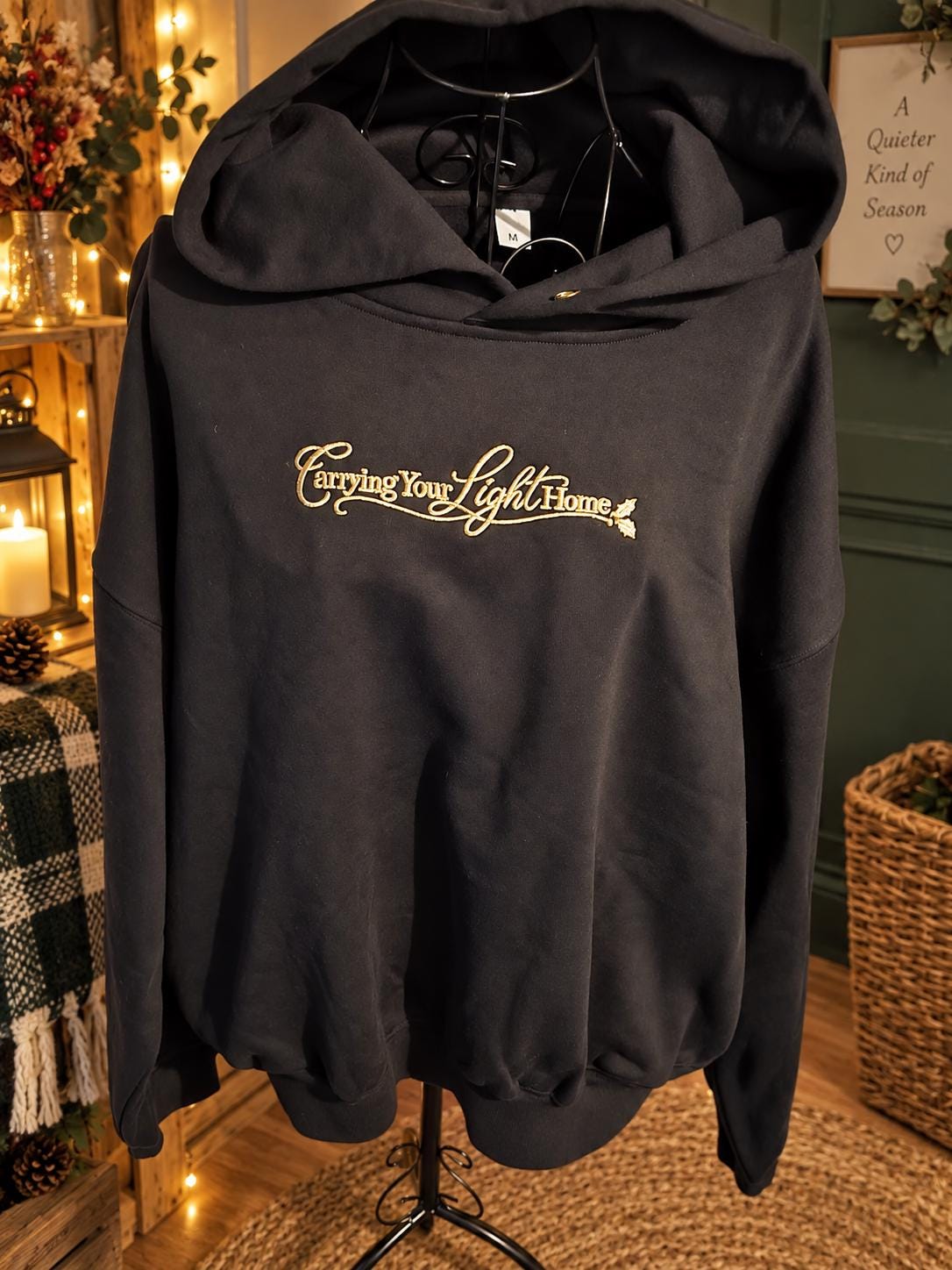 Black heavyweight embroidered hoodie with Carrying Your Light Home gold design in a cosy Christmas setting.
