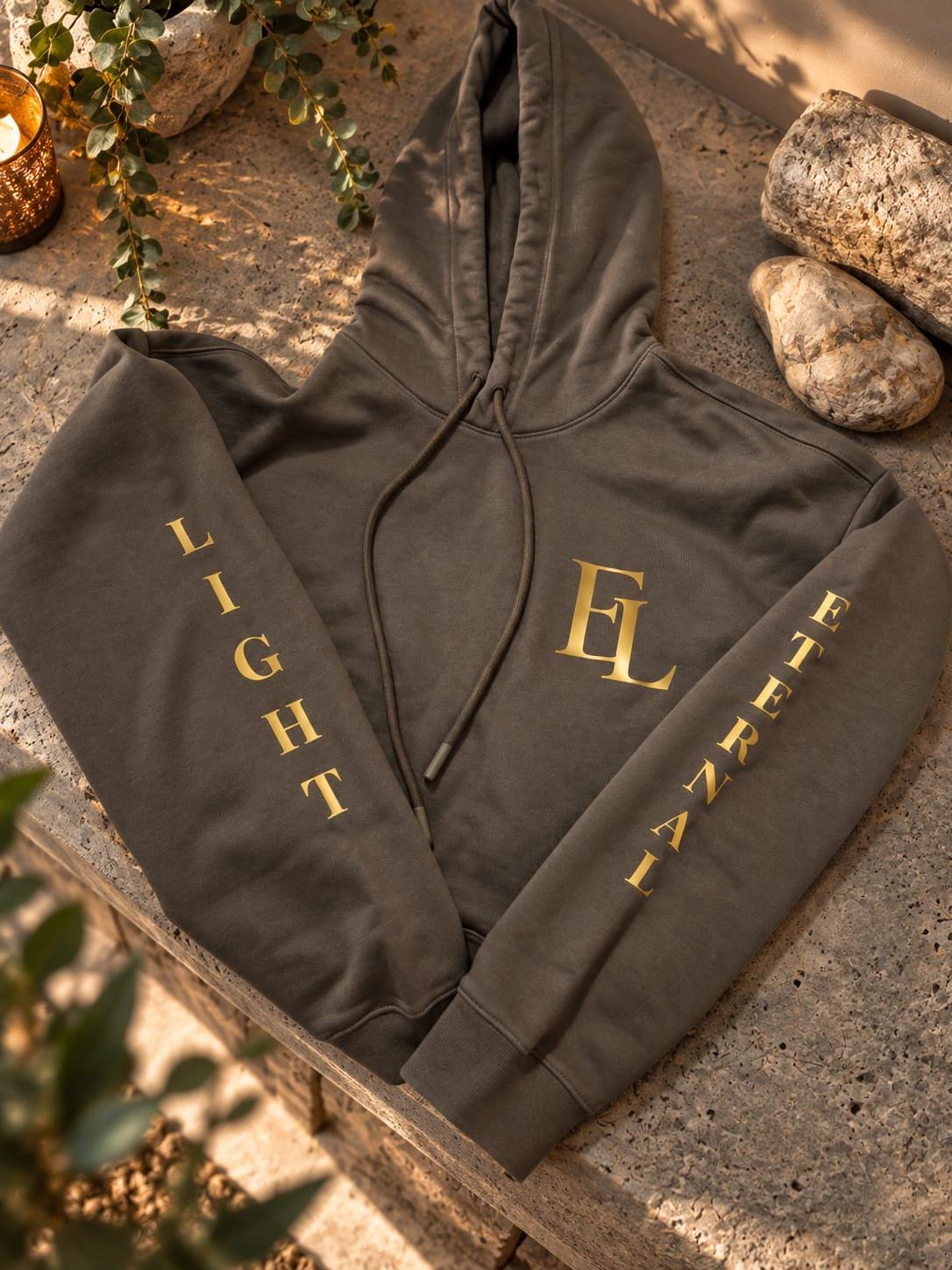 Close view of the charcoal green hoodie laid on stone, with gold LIGHT and ETERNAL lettering running down the sleeves beside the EL monogram.