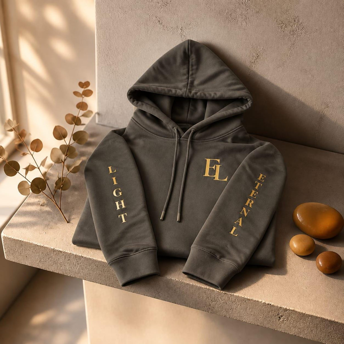 Charcoal green Eternal Light hoodie folded on a pale stone shelf in warm sunlight, showing the gold EL chest monogram and lettering on both sleeves.