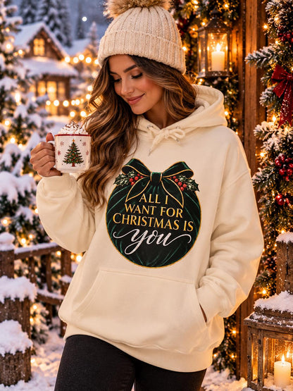 Woman wearing a cream Christmas hoodie featuring an emerald-green and gold “All I Want for Christmas Is You” design, standing outside a cosy festive cabin surrounded by snow, Christmas decorations and warm fairy lights.