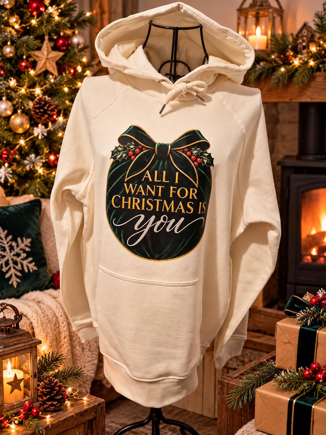 Cream Christmas hoodie displayed on a mannequin in a cosy festive setting with a decorated Christmas tree, glowing fireplace and wrapped gifts, featuring an emerald-green and gold “All I Want for Christmas Is You” design.