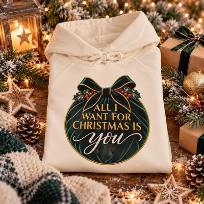 Folded cream Christmas hoodie featuring an elegant emerald-green velvet-effect ornament with gold detailing, festive holly, bow and “All I Want for Christmas Is You” lettering.