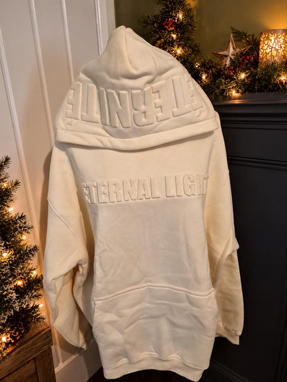 Cream Eternal Light hoodie, showing raised tonal lettering across the front and ÉTERNITÉ detailing on the hood in a cosy Christmas setting.