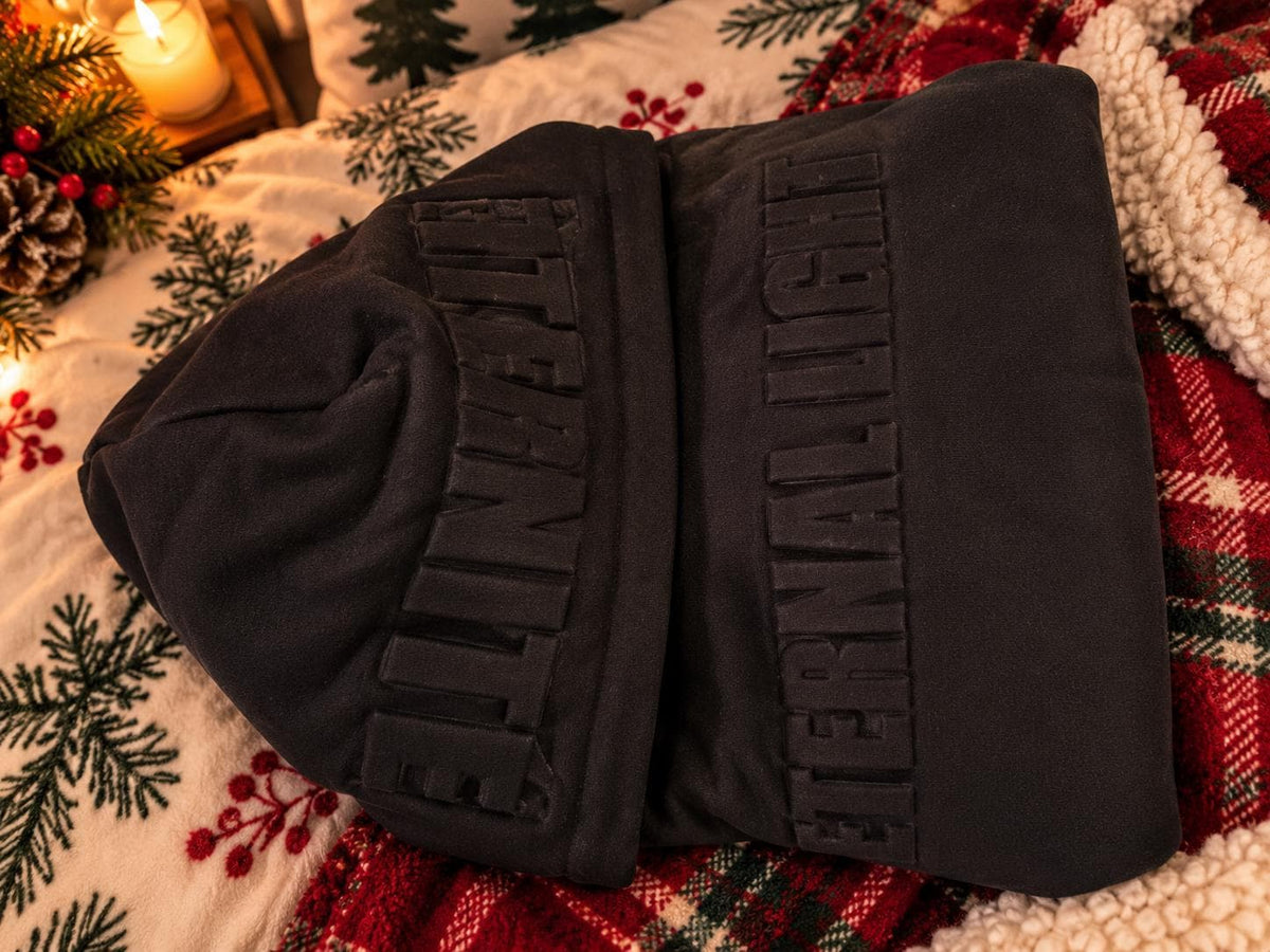 Black Eternal Light hoodie folded sideways on festive bedding, highlighting the raised ÉTERNITÉ hood lettering and soft cream fabric.
