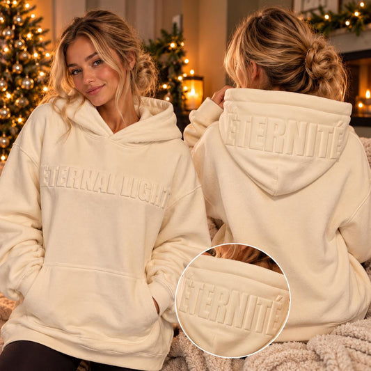 Woman modelling a cream Eternal Light hoodie, showing raised tonal lettering across the front and ÉTERNITÉ detailing on the hood in a cosy Christmas setting.