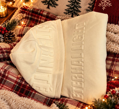 Cream Eternal Light hoodie folded sideways on festive bedding, highlighting the raised ÉTERNITÉ hood lettering and soft cream fabric.