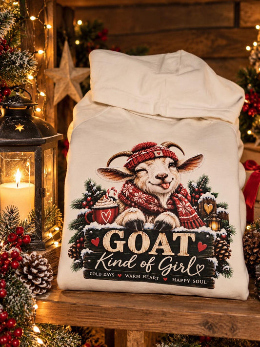 Folded cream GOAT Kind of Girl Christmas hoodie displayed on a rustic wooden shelf beside a glowing lantern, featuring a winking goat in a red festive hat and scarf, surrounded by Christmas greenery, pinecones and warm fairy lights.