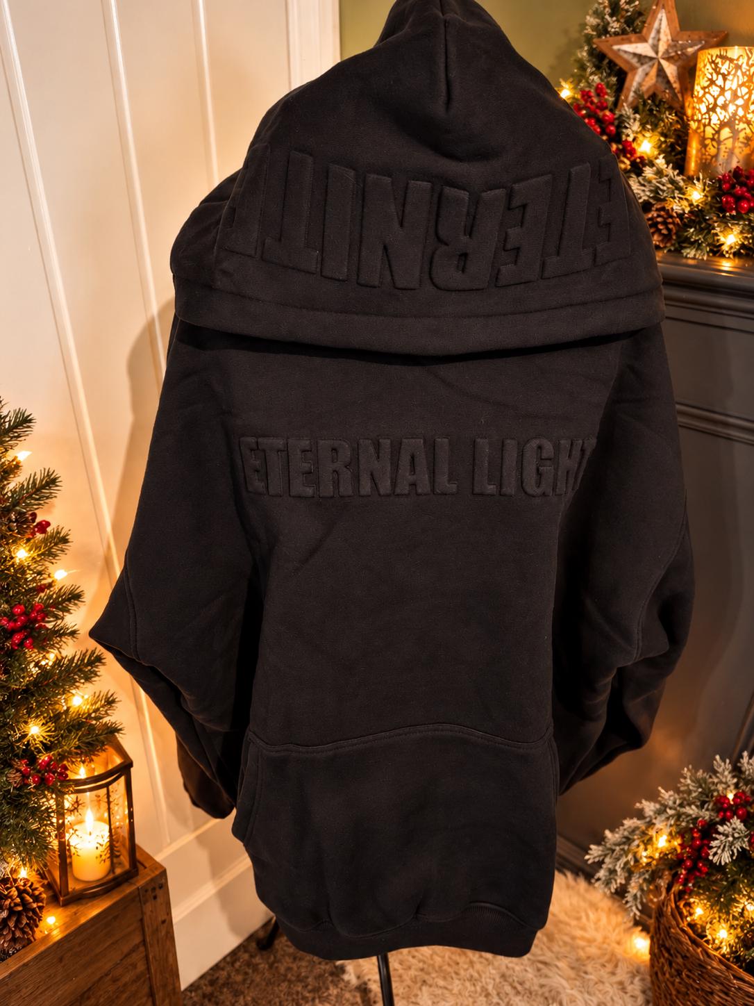 Black Eternal Light hoodie, showing raised tonal lettering across the front and ÉTERNITÉ detailing on the hood in a cosy Christmas setting.